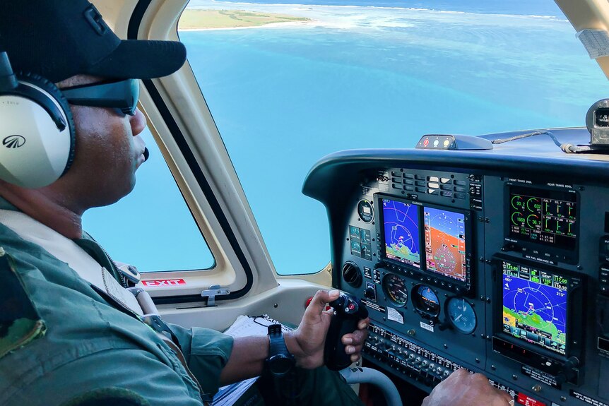 A man wearing a cap and headphones steers a plane as it circle above an island shore.