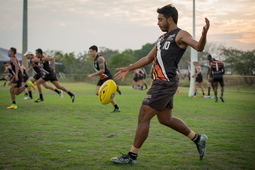A young Indigenous footballer kicks a football as his teammates run behind him in the background.