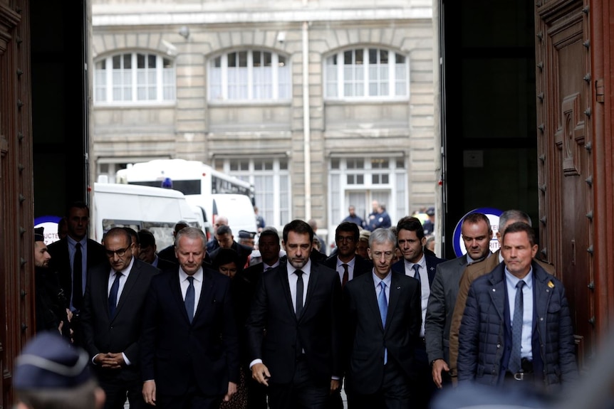 Christophe Castaner walks with a group of other men in suits through the gate of a walled courtyard with police vehicles.