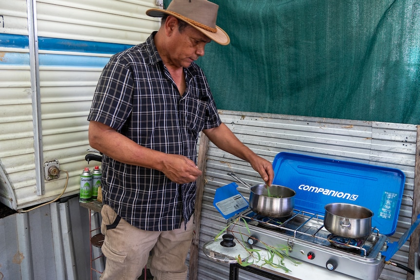 Aboriginal man puts leaves into pot on camp stove