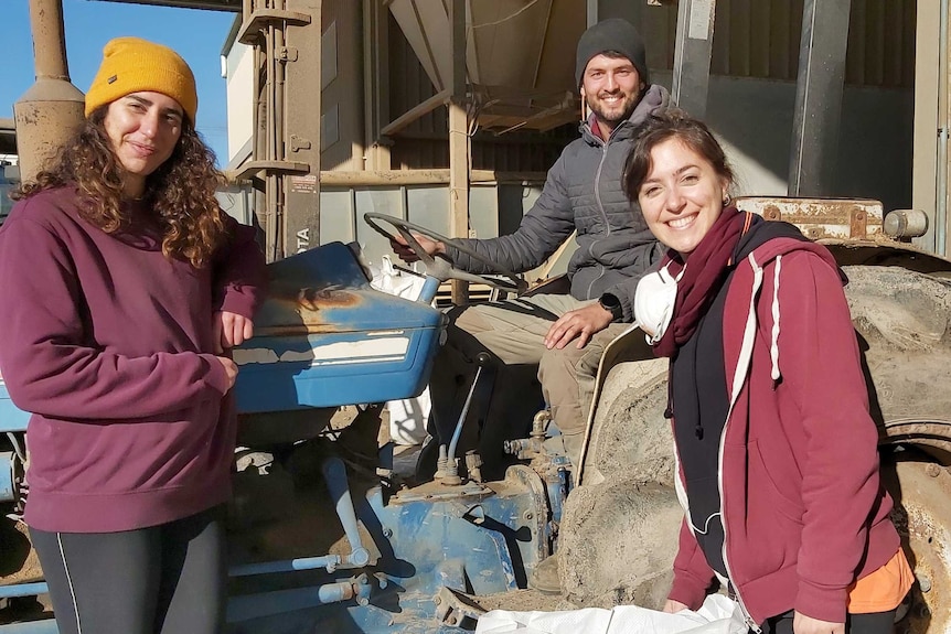 A young man sits in an old tractor and two females stand in front outside a seed warehouse
