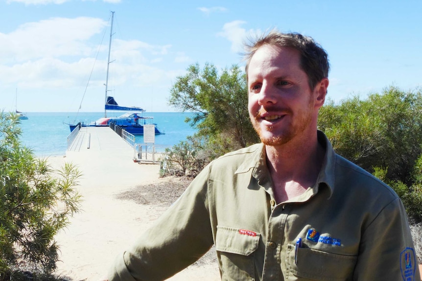 A man in a khaki shirt stands in front of a jetty with a yacht moored in light blue waters.