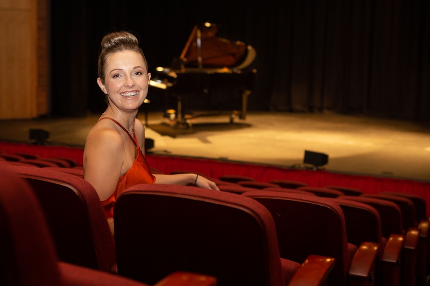 A woman in evening gown and hair ready for the stage sits in the theatre seating, stage behind.