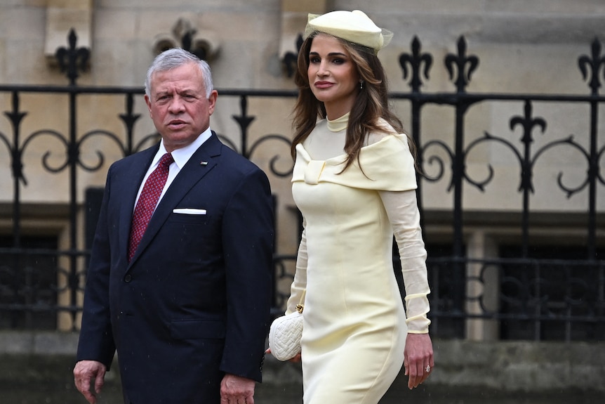 Jordan's King Abdullah II and Queen Rania walking in formal wear beside a fence.