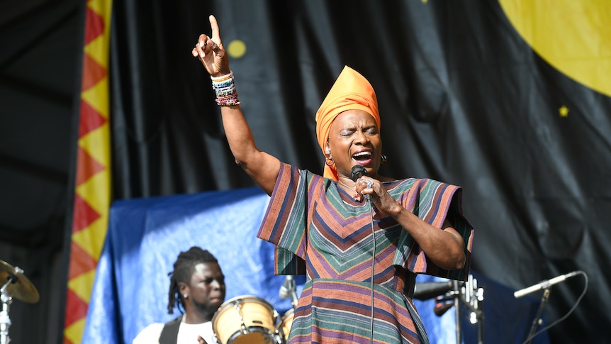 Angelique sings into a mic on a festival stage, one hand pointing in the air. She is wearing a bright orange patterned outfit