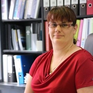 A professional image of a woman with glasses and a fringe in a red blouse, sitting inside an office with files behind her.
