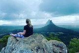 The back of a girl, sitting on the summit, looking out over the Glass House Mountains.