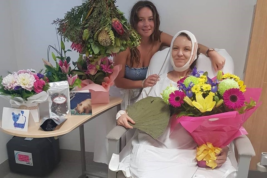 A woman wearing hospital gowning sits with her head bandaged as a teenager embraces her