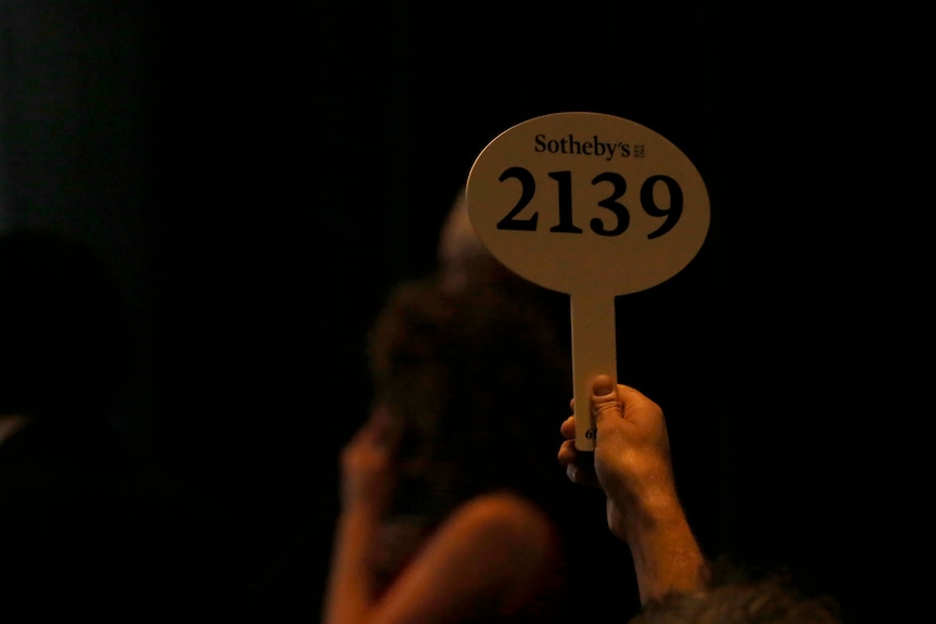 A hand holds up an oval-shaped bidding paddle with the numbers '2139' on it and the Sotheby's logo at the top in a dim room.