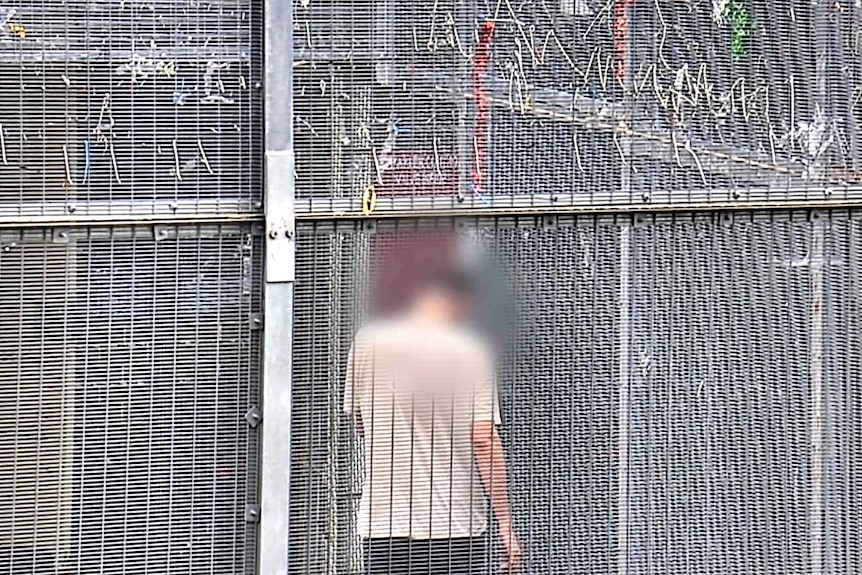 A man's back and blurred head can be seen through a high-security tall fence in CCTV footage.