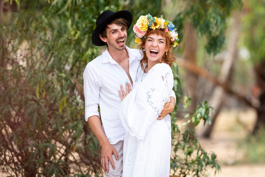 A couple dressed in white laugh in front of trees. 
