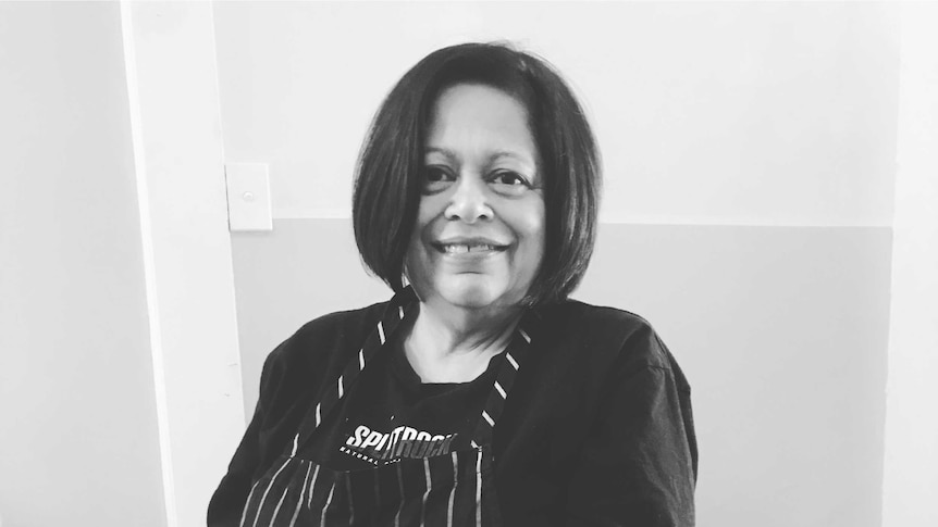 Black and white photo of author and chef Ragini Dey in a striped apron holding a large pot of lentils