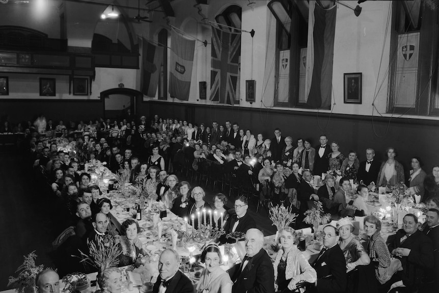 Black and white photo showing long tables of men and women in evening dress at a lavish dinner.