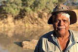 A close up photo shows a the face of a man wearing a hat in the late afternoon sun, with a creek, creek bank and shrubs behind.