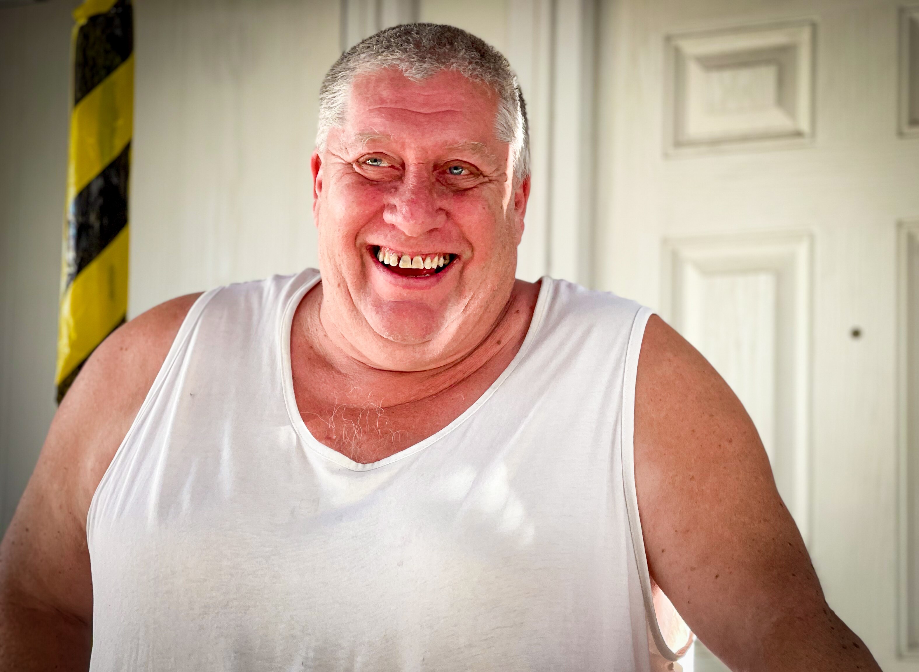 A man with short grey hair in a white singlet smiles while stand out front of his house.