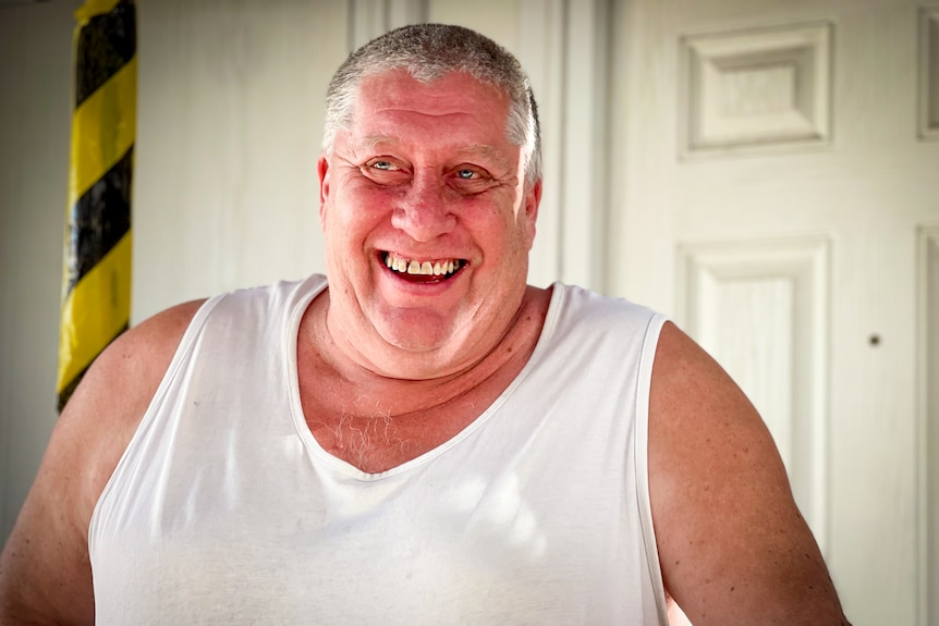A man with short grey hair in a white singlet smiles while stand out front of his house.