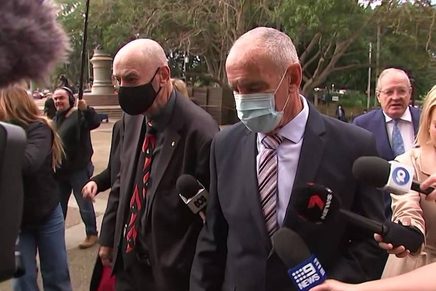 Two men wearing masks walk in a crowd of media