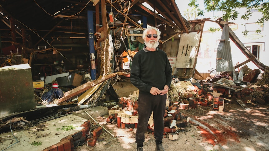 Man in black pants and jumper, sunglasses, stands in front of a severely damaged mechanic's garage