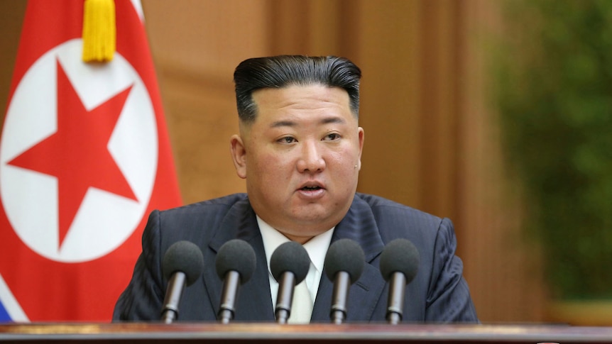 North Korean leader Kim Jong Un delivers a speech seated at a desk with  an array of microphones