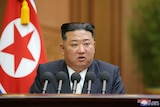 North Korean leader Kim Jong Un delivers a speech seated at a desk with  an array of microphones