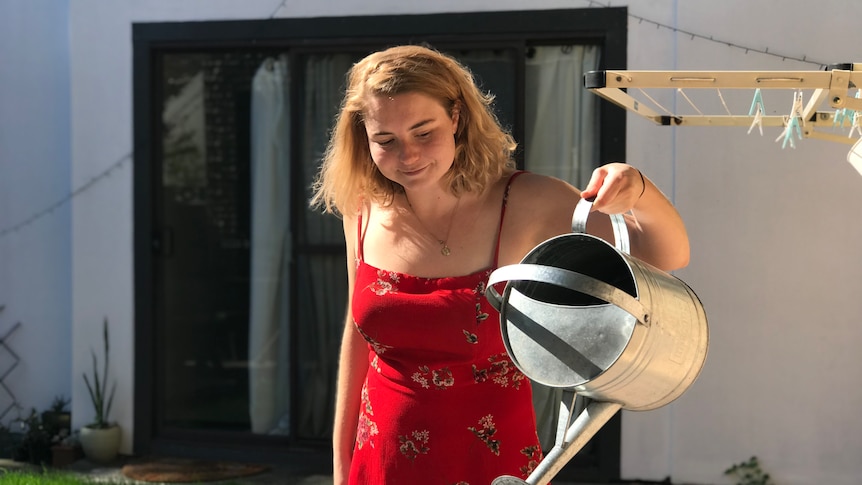 A woman in a red dress looking down as she waters her garden with a silver watering can. 