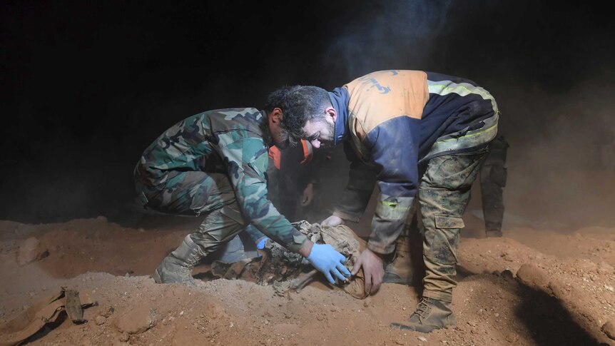 Two men carefully collect human bones from a dusty mass grave.
