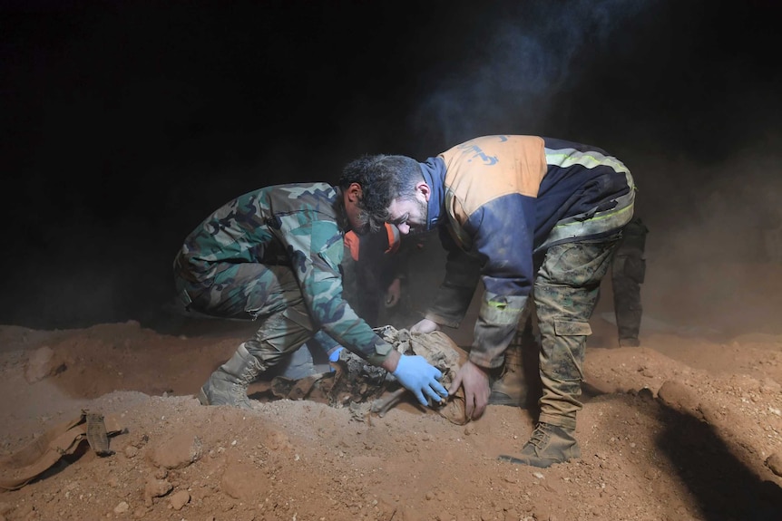 Two men carefully collect human bones from a dusty mass grave.