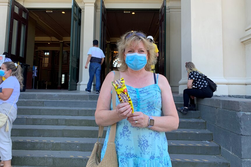 A woman in a blue dress and a blue surgical mask holds up an ice cream on a sunny day.