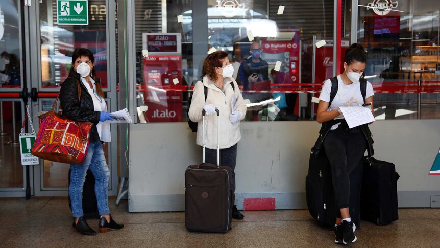Three woman wearing white masks and plastic gloves wait with bags and suitcases at a train platform in Italy