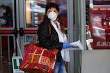 Three woman wearing white masks and plastic gloves wait with bags and suitcases at a train platform in Italy