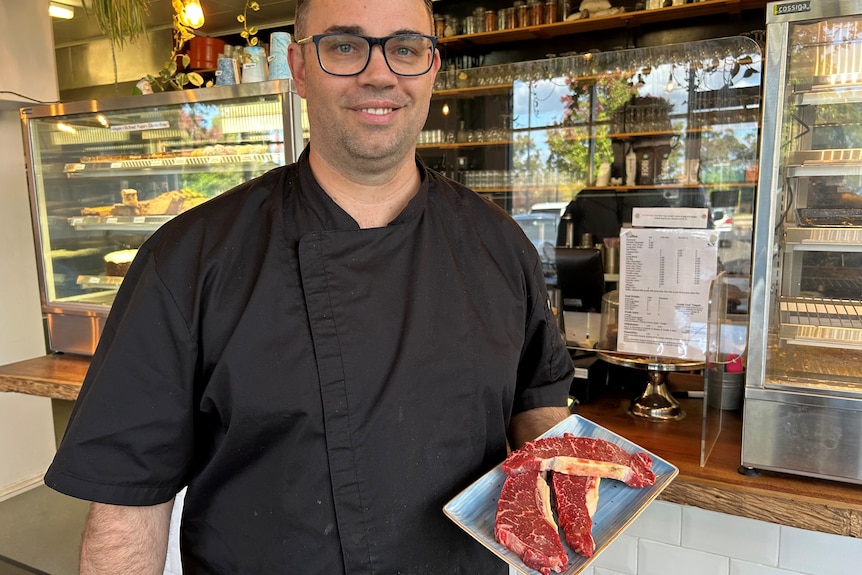 A man wearing a black chef uniform holds a plate with three steaks on it.