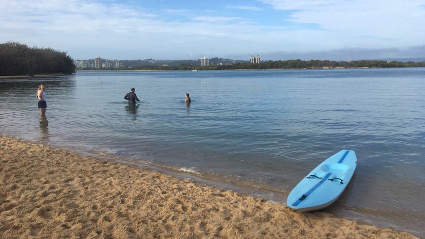 A man and two girls stand in the still river water with a kayak on the beach in the foreground.