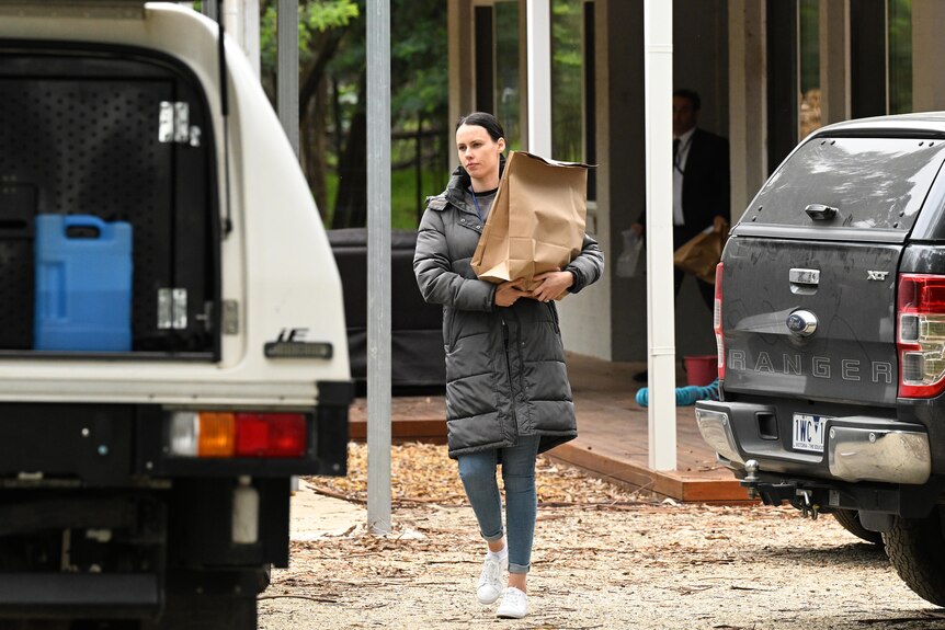 A policewoman walks between two cars carrying a brown paper evidence bag.