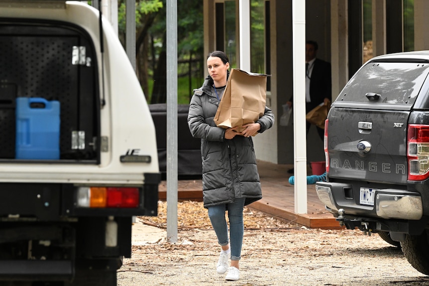A policewoman walks between two cars carrying a brown paper evidence bag.