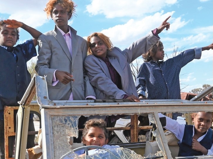 Six young Indigenous boys wearing men's suits pose crowded in car with broken windscreen