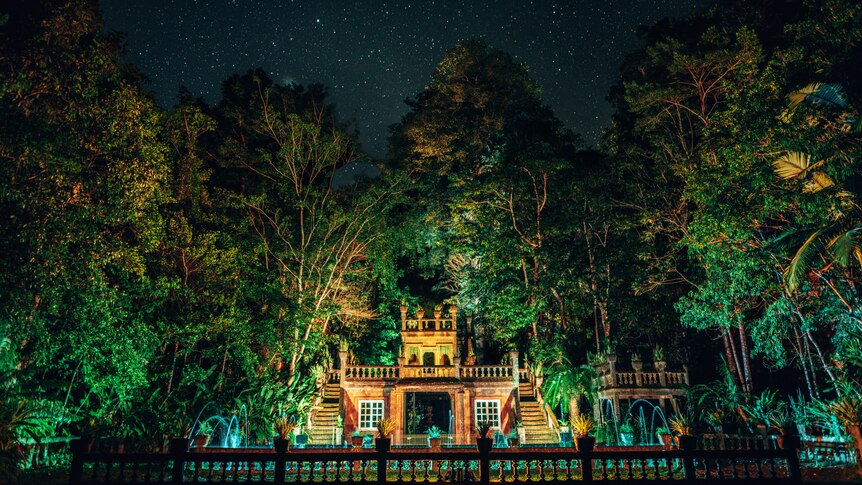 A photo of a castle at night in a forest, with everything all lit up by lights.