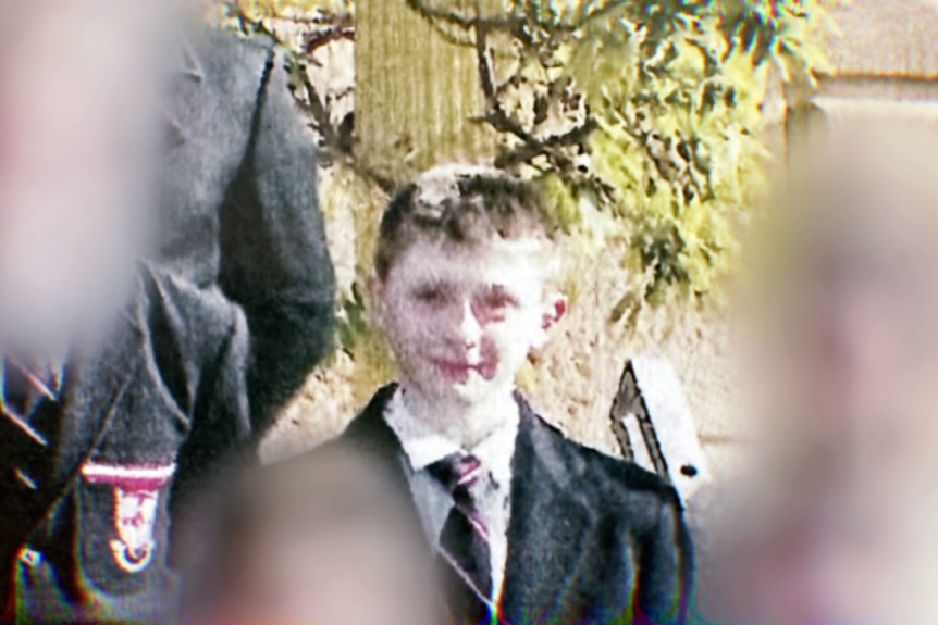 A grainy photo of a young boy in uniform posing for a school photograph with other student faces blurred.