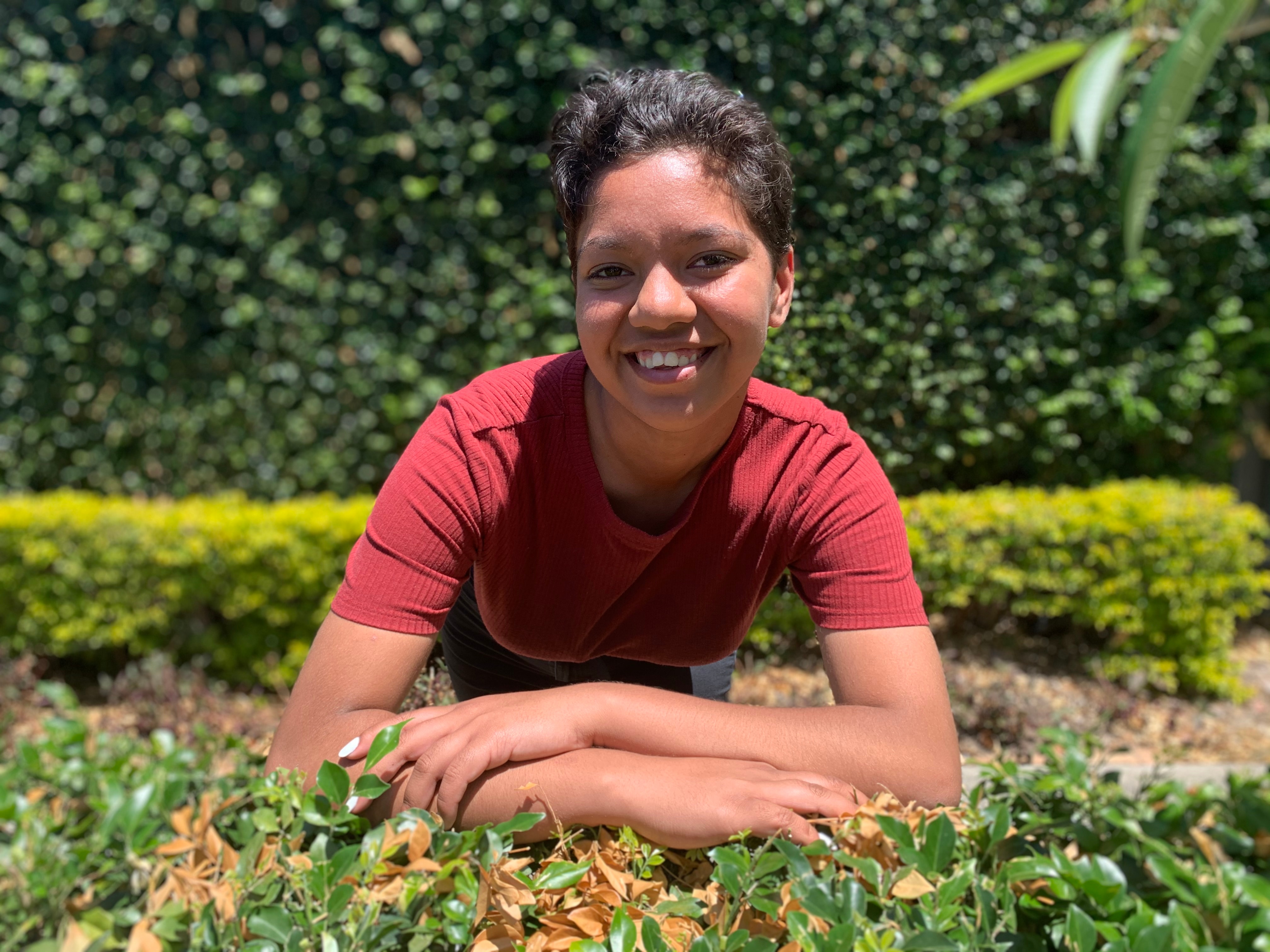 A young woman leans onto a hedge as she looks to the camera and smiles. She wears a red top and has short hair