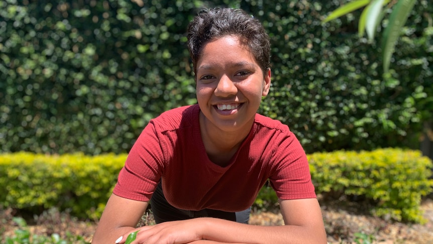 A young woman leans onto a hedge as she looks to the camera and smiles. She wears a red top and has short hair