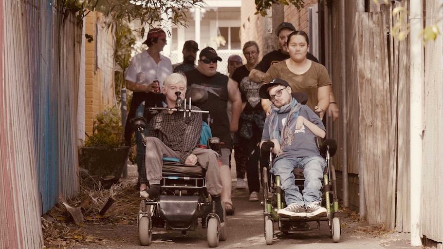 A group of people, including two people in wheelchairs, walk down an alleyway while filming a scene for music video.