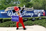 A person wearing a mouse costume dances in front of Disney park sign as other put up poster with words 'Pedo world'.