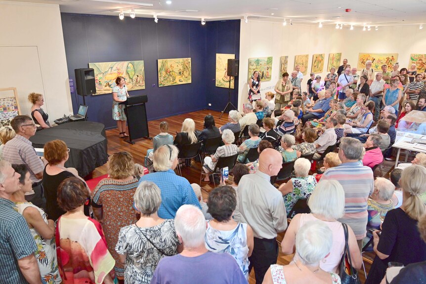 A large crowd listening to a speaker inside an art gallery, with paintings on the walls.