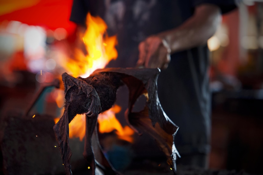 The roasted carcass of a bat is held on a stick, with flames visible in the background