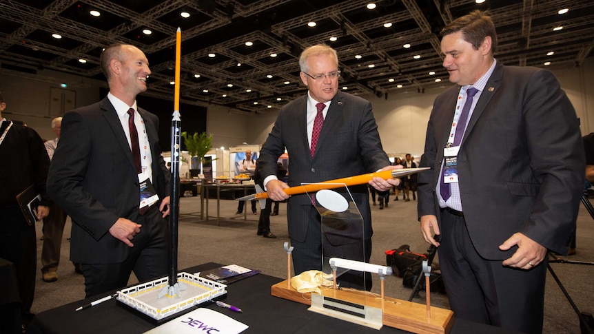 Three men in suits stand in a large room around a table, the man in the centre is holding a yellow model rocket.