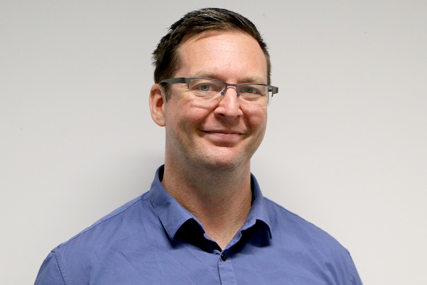 Un homme en chemise bleue avec des cheveux noirs et des lunettes sourit à la caméra. 