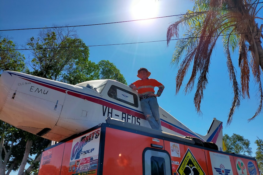 Young man wearing bright orange shirt and jeans stands atop an orange caravan, leaning against a light aircraft.