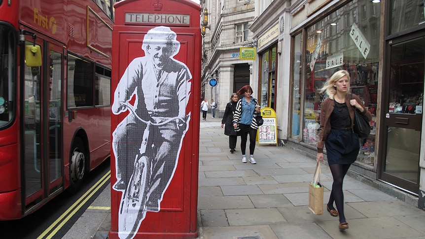 A photo of street art of Albert Einstein on a bike on a red London Phone Booth on the street. A woman is walking past.