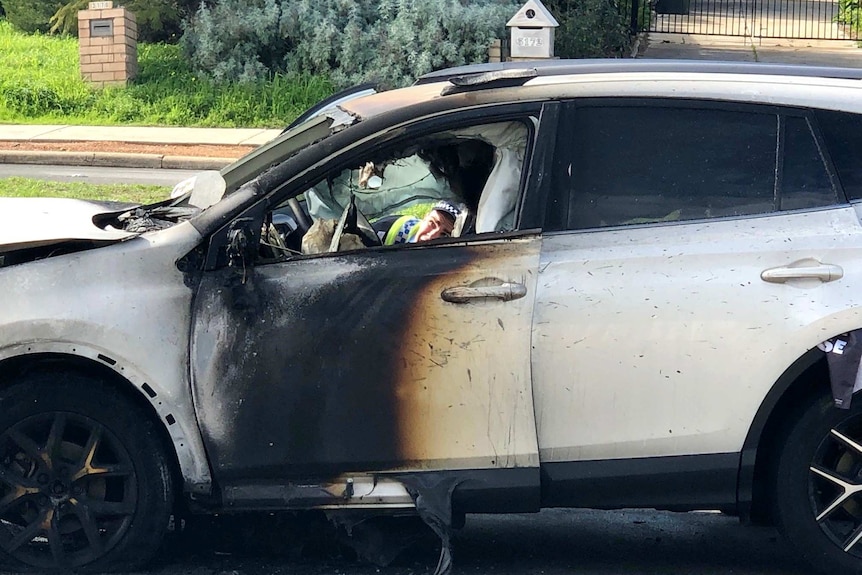 A close up shot of a police officer peering through the driver's side door of a burnt out car.