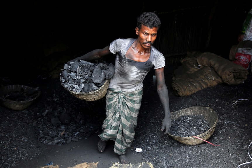 A young Indian man with a moustache in muddied T-shirt and sarong carries a basket of coal in mine