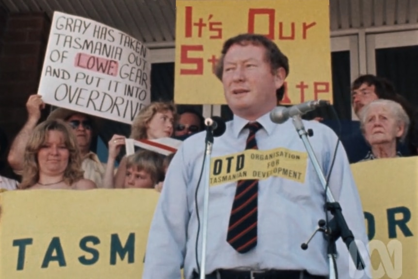 A man in a blue shirt addresses a crowd in front of signs reading "It's our state".